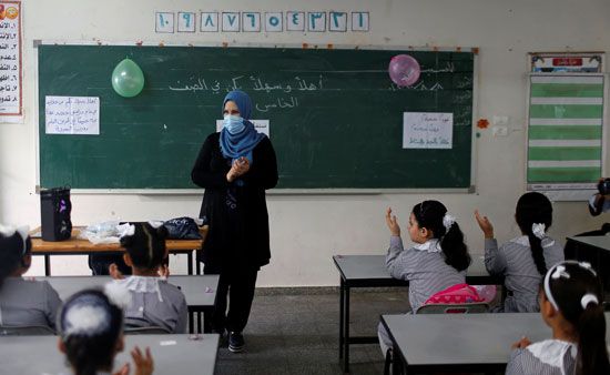 مدرسة ترتدي قناع وجه واقي يشير بينما يجلس الطلاب الفلسطينيون في فصل دراسي